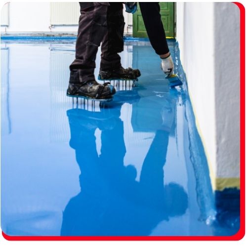 Worker applying blue epoxy flooring along the edge with spiked shoes in an Aurora facility.