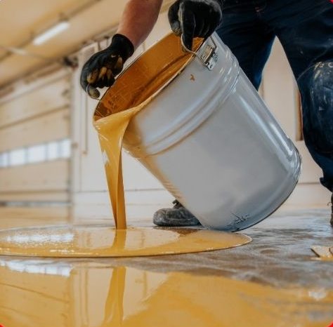 Worker pours yellow epoxy coating from a metal bucket onto a floor in a Hamilton industrial setting.