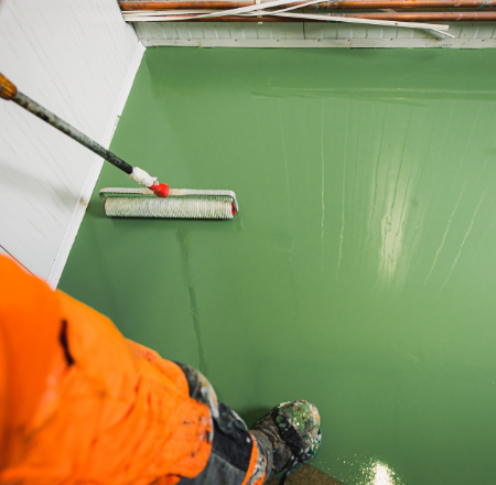Worker in orange gear applies green epoxy coating with a roller in Muskoka facility.